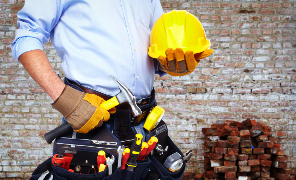 A worker holding a yellow hard hat and wearing a tool belt in front of a brick wall