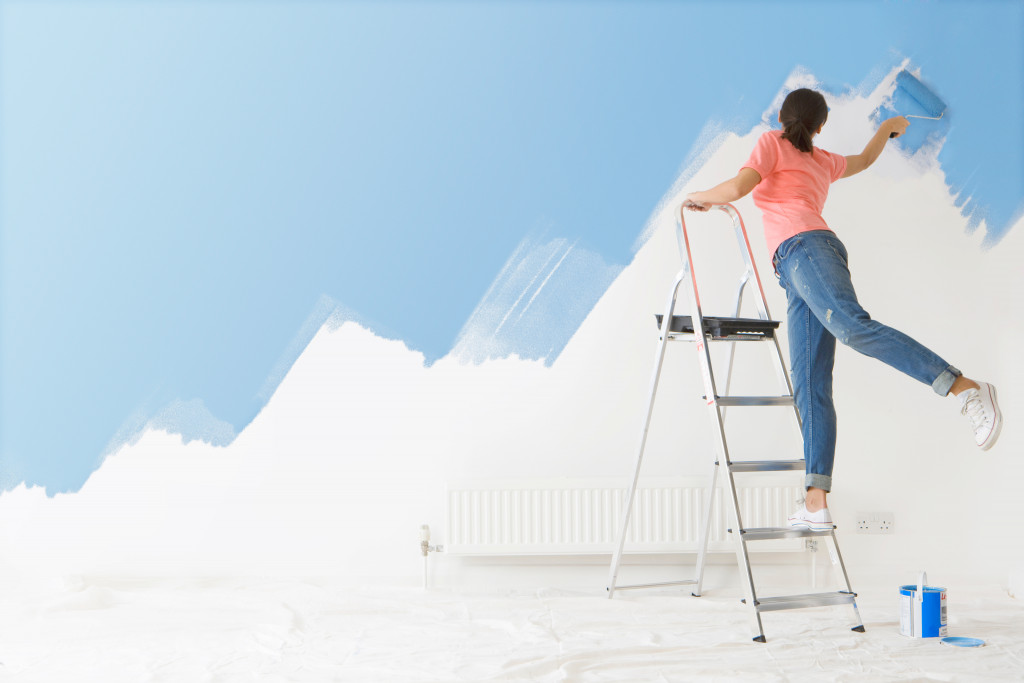 woman painting white wall blue while on top of step ladder