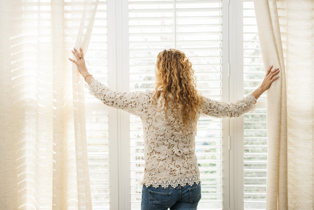 woman letting in natural light by opening up light-colored curtains in windows